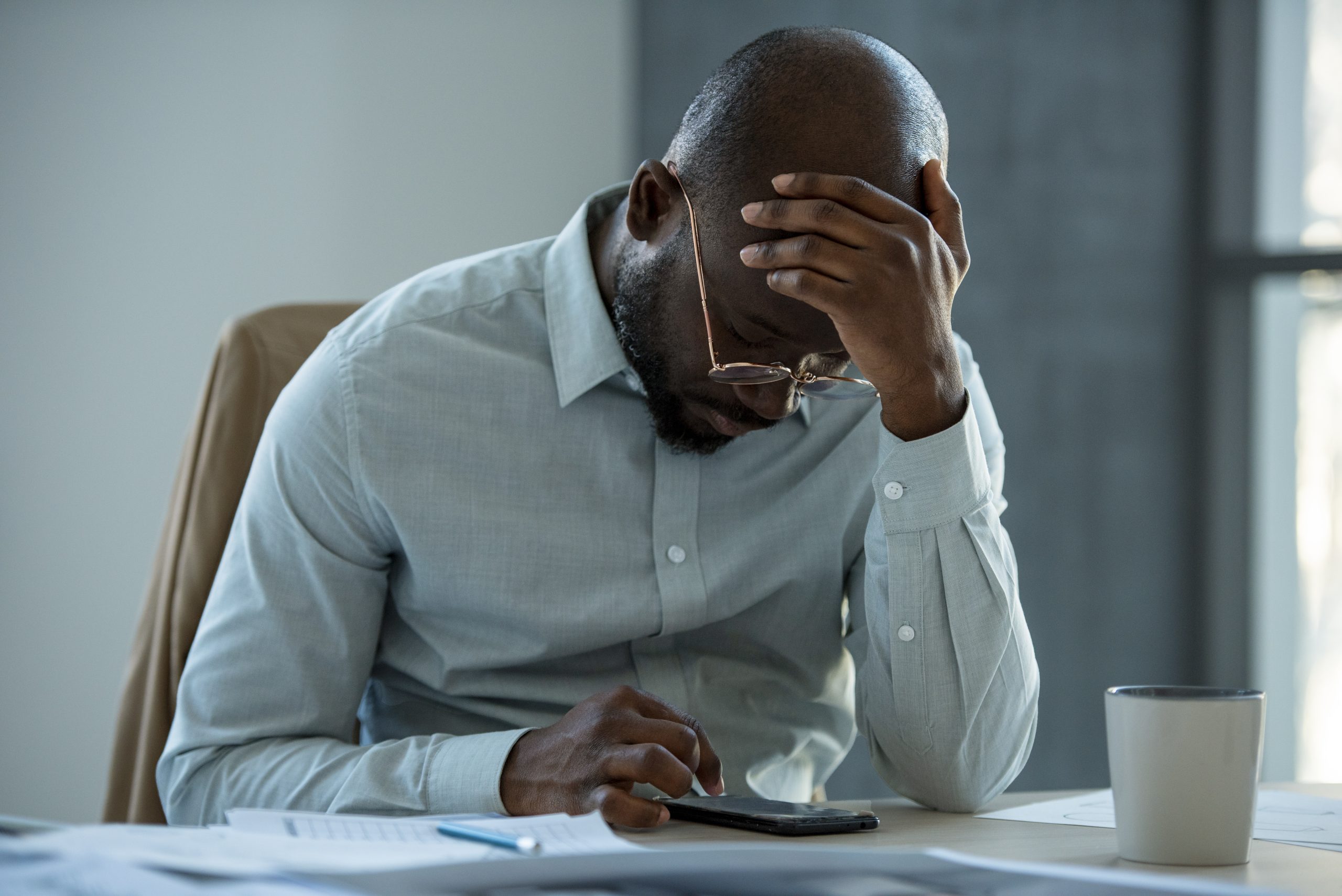 Homem negro de meia-idade, vestindo camisa social clara, sentado à mesa de um escritório com expressão de preocupação. Ele segura um celular com uma das mãos enquanto apoia a cabeça na outra, parecendo estressado ou com ansiedade financeira. A mesa contém papéis, uma caneta e uma xícara, sugerindo um ambiente de trabalho.