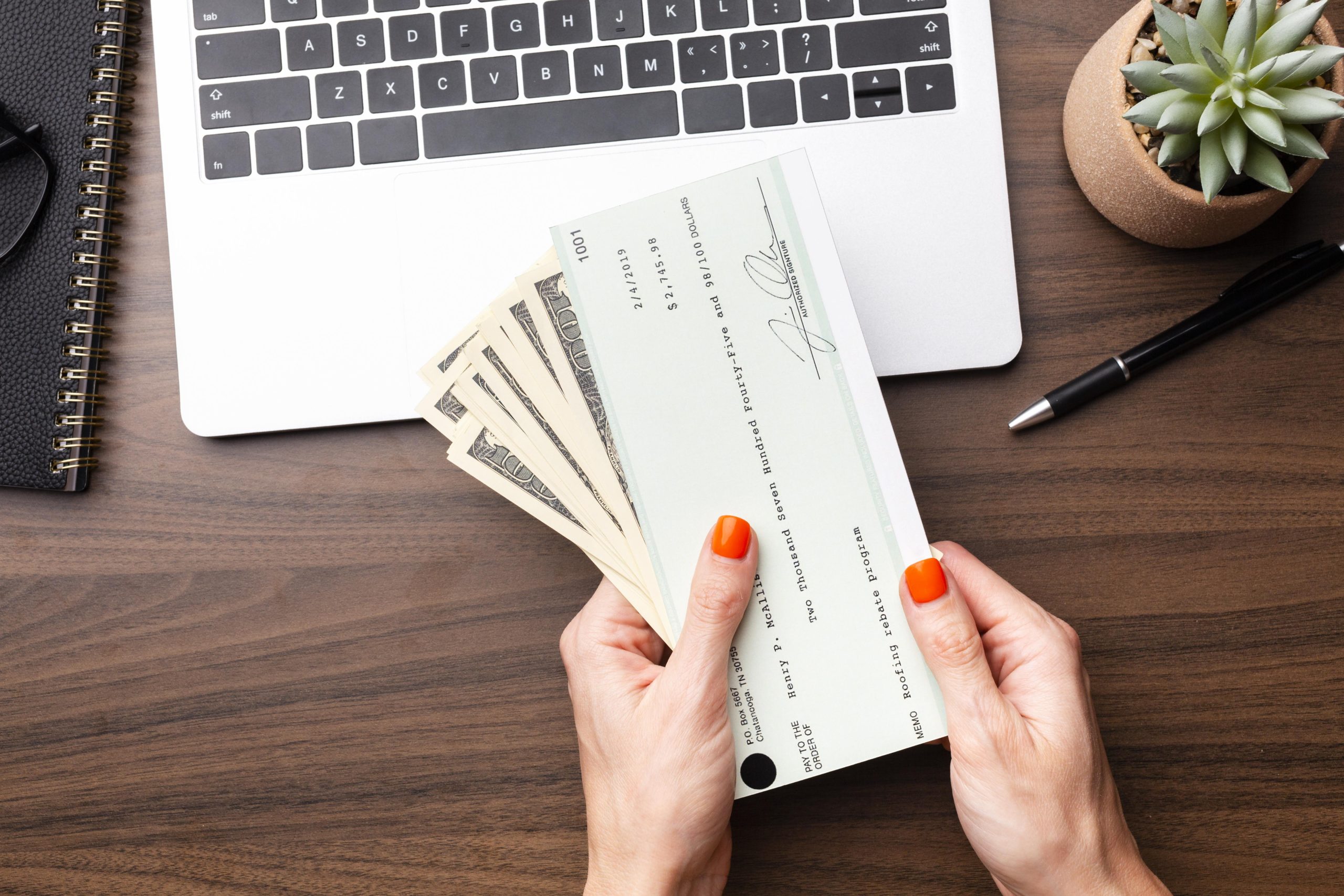 Mãos femininas com unhas pintadas seguram um cheque e várias notas de dólar sobre uma mesa de escritório, ao lado de um laptop, um caderno e uma caneta. A cena sugere uma transação financeira, pagamento ou recebimento de valores, destacando a importância da gestão financeira e do uso de meios tradicionais de pagamento.