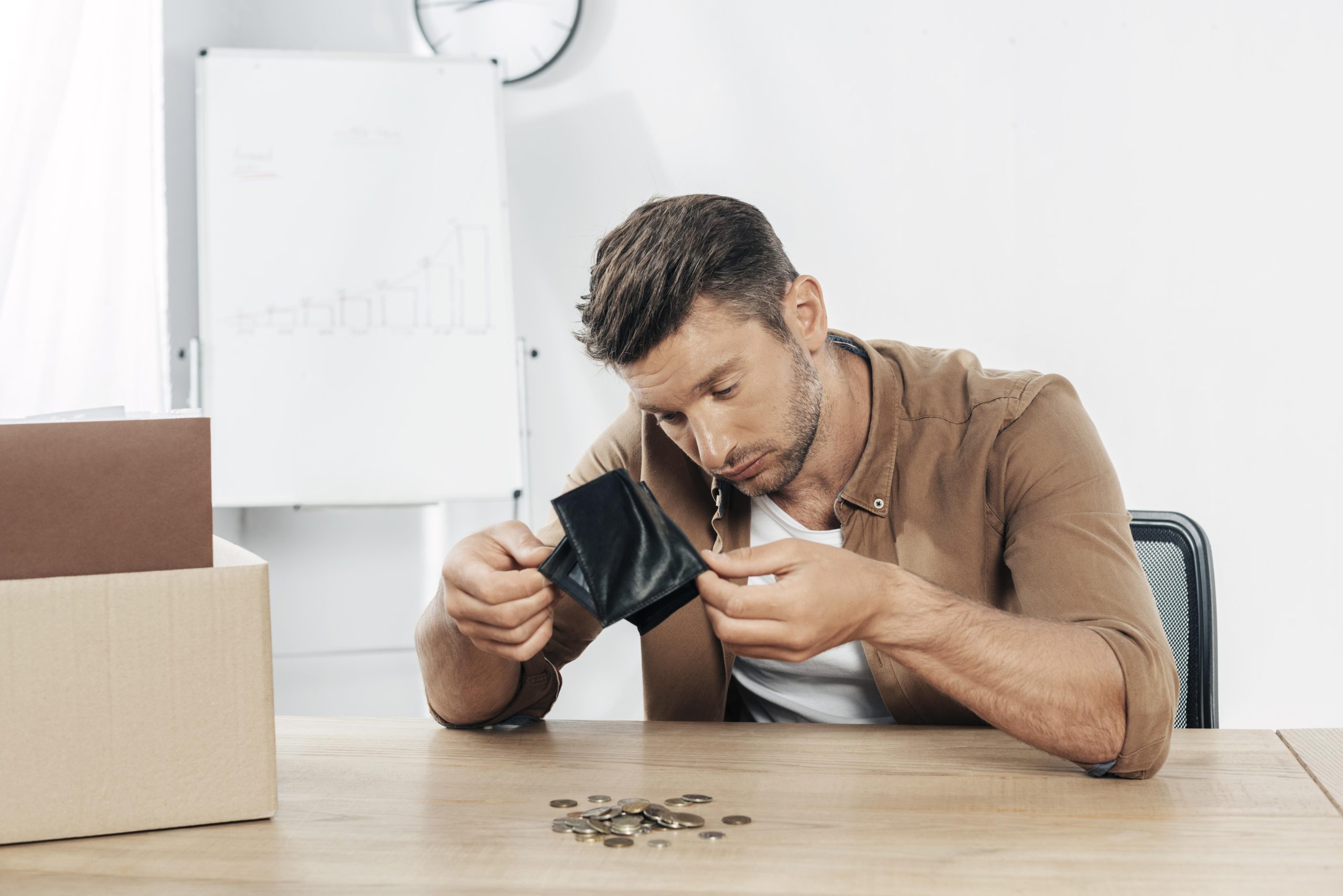 Homem jovem de aparência preocupada sentado à mesa de um escritório, segurando uma carteira vazia enquanto observa um pequeno montante de moedas espalhadas à sua frente, simbolizando dificuldades financeiras. Ao fundo, um quadro branco com gráficos e um relógio reforçam o ambiente corporativo. A imagem transmite a importância de revisar comportamentos financeiros para evitar crises e melhorar a gestão do dinheiro.
