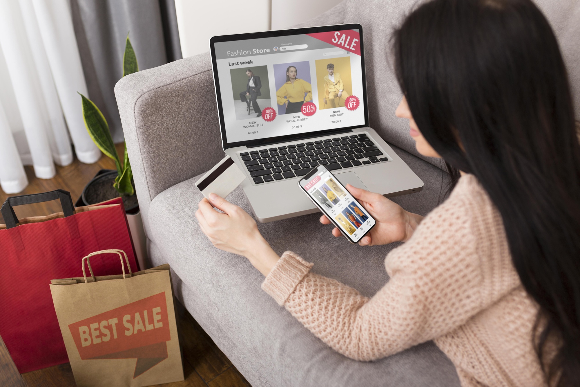 Mulher sentada no sofá segurando cartão de crédito e celular enquanto realiza compras online em sites de roupas, com laptop aberto e sacolas ao lado.