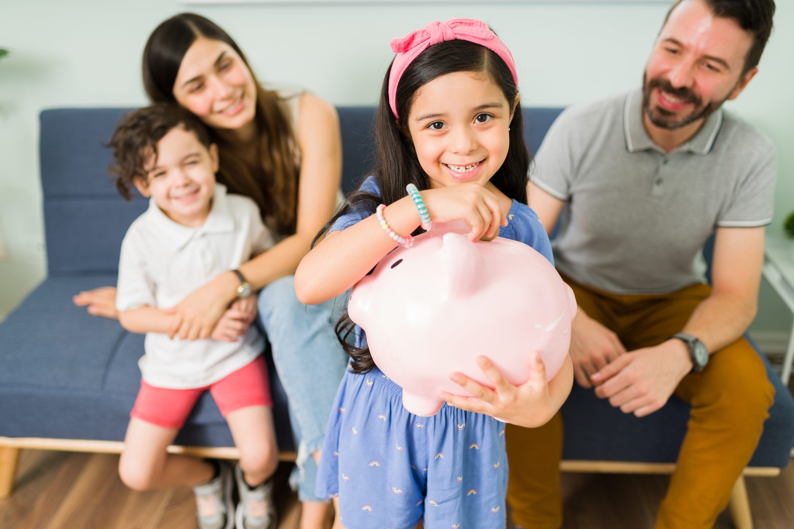 Menina sorridente coloca uma moeda em um cofrinho rosa enquanto seus pais e irmão a observam felizes no sofá, representando um momento de aprendizado sobre educação financeira em família.