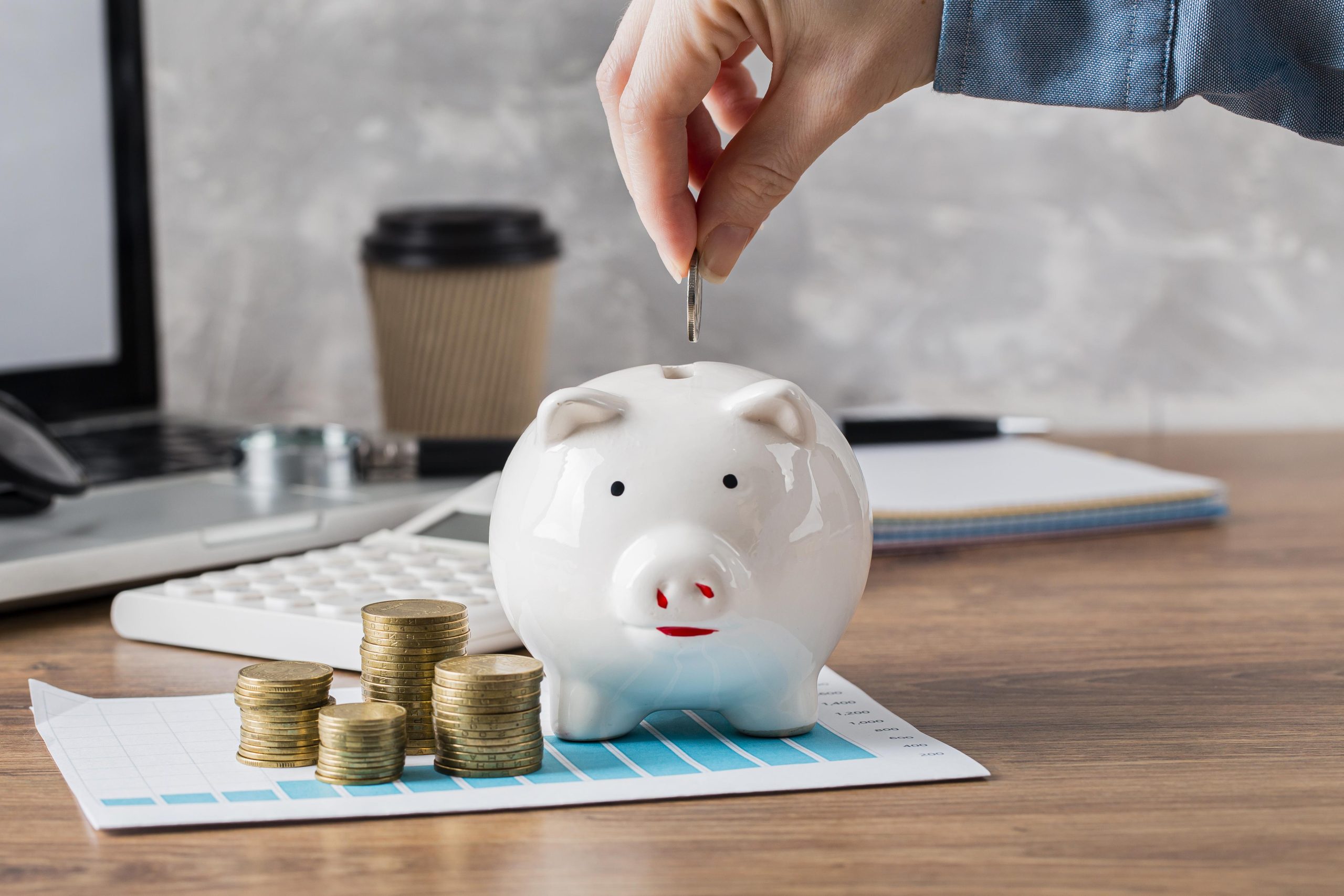 Mão feminina depositando uma moeda em um cofrinho de porcelana branca, simbolizando o hábito de economizar dinheiro. Ao redor, há pilhas de moedas, uma calculadora, um laptop e cadernos sobre a mesa, remetendo à importância da educação financeira para o planejamento e controle dos gastos.