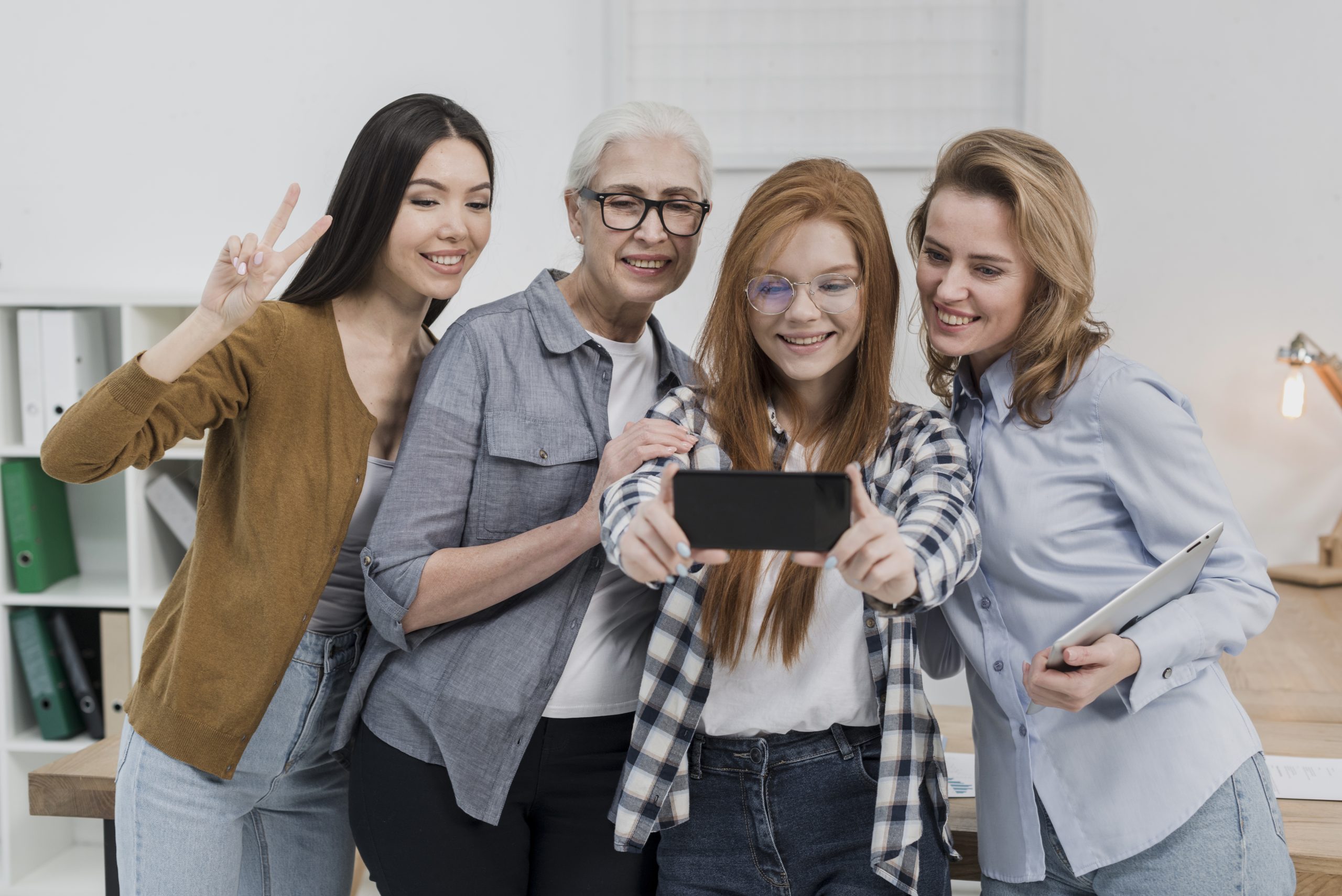 Mulheres de diferentes gerações sorrindo enquanto tiram uma selfie juntas em um ambiente de escritório, representando a conexão entre idades na hora de aprender e escolher produtos financeiros.