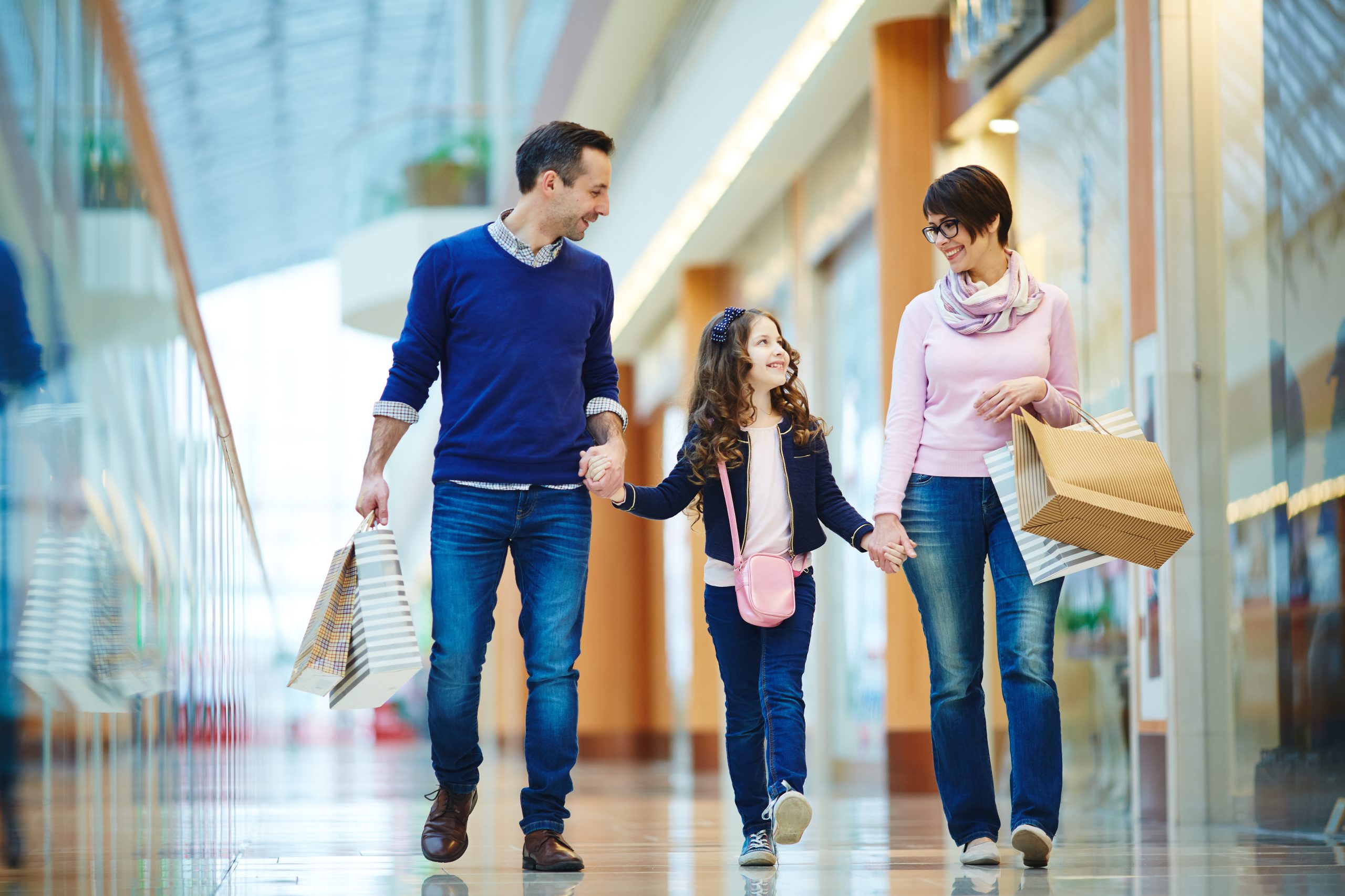 Família sorridente da classe média no Brasil caminha junta em um shopping center moderno. O pai e a mãe seguram sacolas de compras enquanto acompanham a filha pequena, que caminha alegre entre eles. A imagem transmite conforto, consumo consciente e bem-estar típico da rotina de lazer da classe média no Brasil.