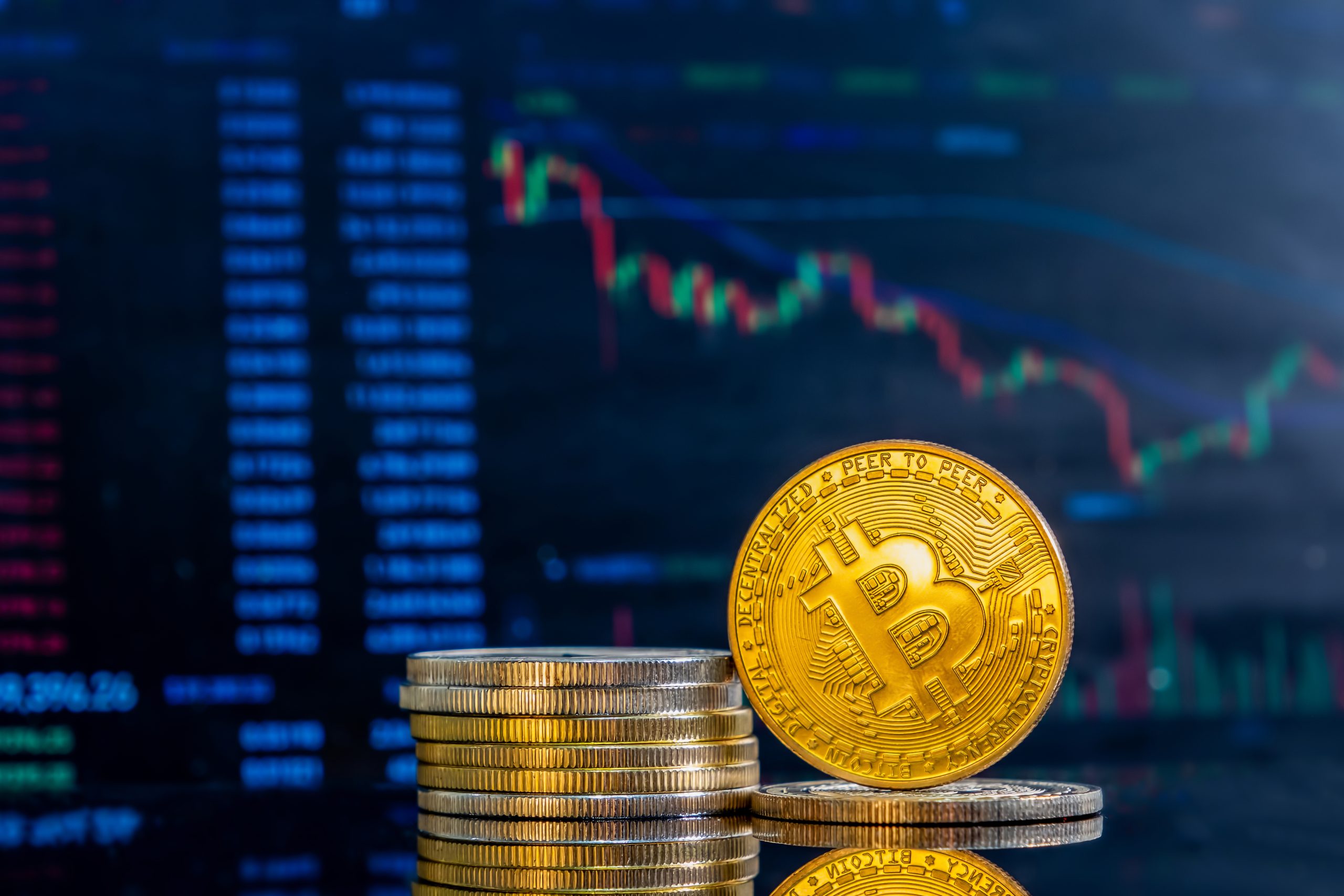 Várias moedas empilhadas com uma moeda dourada de Bitcoin em destaque à frente, representando o conceito de criptomoeda. Ao fundo, uma tela desfocada exibe gráficos financeiros coloridos e dados de mercado, ilustrando a volatilidade e as tendências de investimento em criptomoedas. A composição reforça a ideia de inovação tecnológica e finanças digitais.