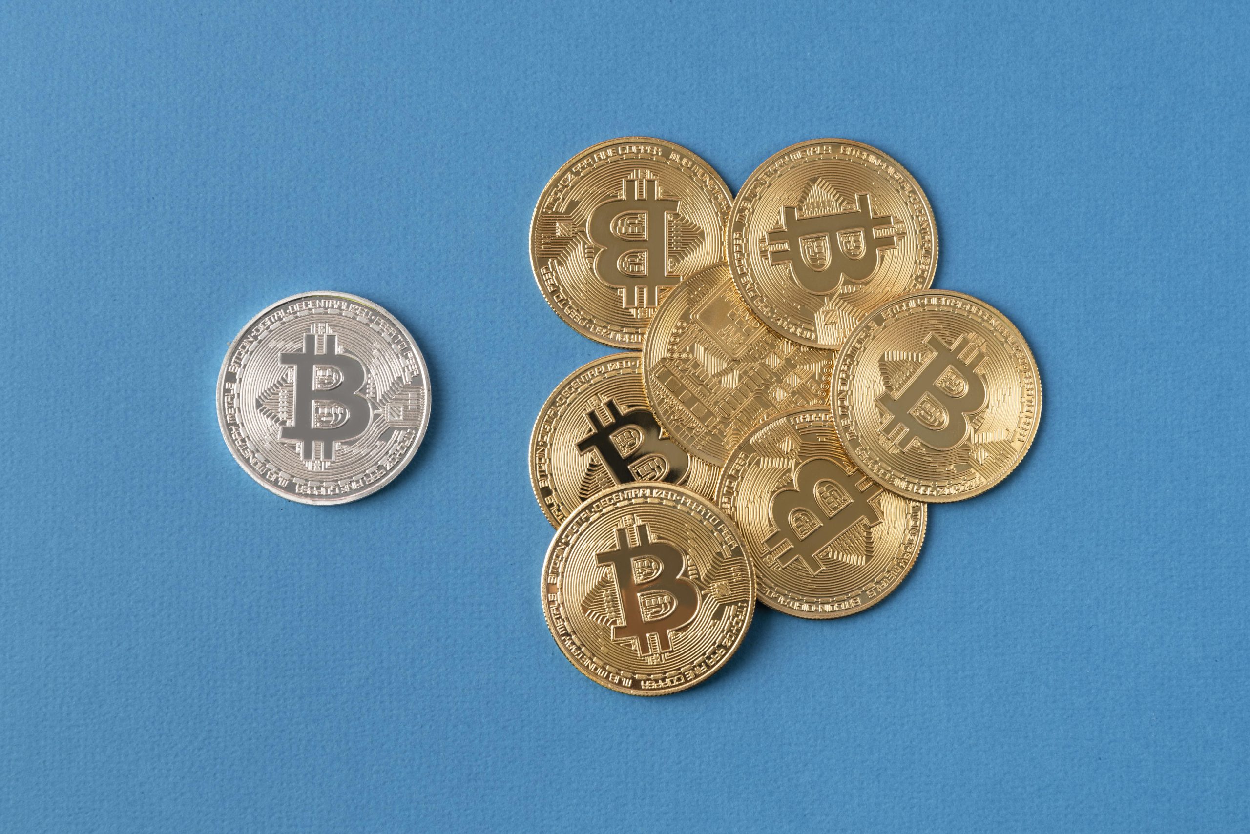 Diversas criptomoedas representadas por moedas físicas douradas e uma prateada, todas com o símbolo do Bitcoin, dispostas sobre uma superfície azul vibrante.