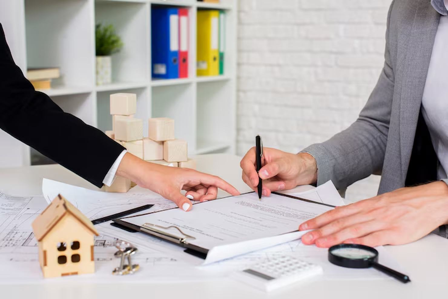 Duas pessoas analisam e assinam papéis sobre uma mesa com miniatura de casa, calculadora e plantas arquitetônicas — uma representação simbólica dos documentos para compra e venda de imóvel, fundamentais para garantir segurança e legalidade na transação.