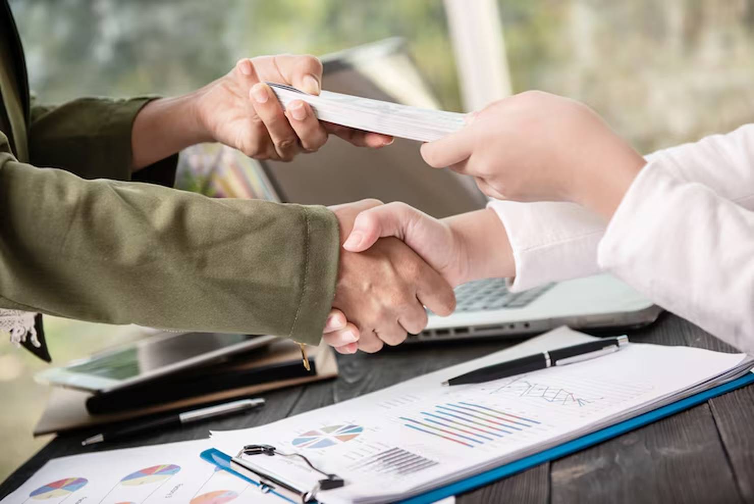 Duas pessoas apertam as mãos enquanto uma delas entrega um maço de dinheiro, em um ambiente de trabalho com gráficos financeiros sobre a mesa — simbolizando um acordo transparente e responsável relacionado a empréstimo pessoal.