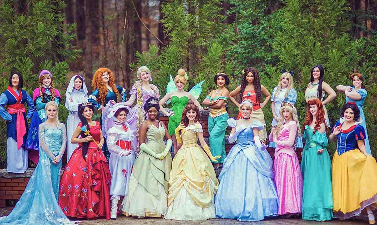 Grupo de mulheres caracterizadas como princesas da Disney, reunidas ao ar livre em frente a uma fileira de árvores, vestindo trajes icônicos de personagens como Cinderela, Ariel, Bela, Elsa, Mulan e outras.