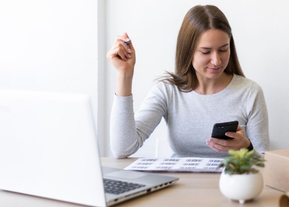 Mulher jovem sentada à mesa de trabalho, olhando o celular com papéis e notebook ao lado, representando a organização financeira e a busca por crédito pessoal de forma prática e segura.