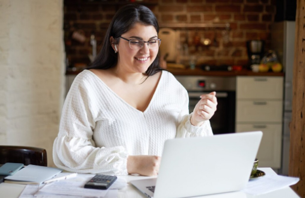 Mulher sorridente usando notebook em uma cozinha moderna, analisando documentos e calculadora ao lado, representando o planejamento financeiro para solicitar um empréstimo pessoal online com segurança.
