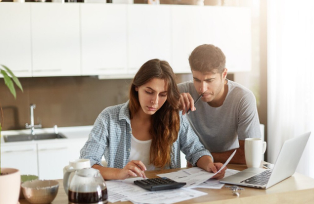 Casal concentrado analisando contas e calculadora em uma mesa com notebook e papéis, simbolizando o planejamento de finanças pessoais e a organização do orçamento doméstico.
