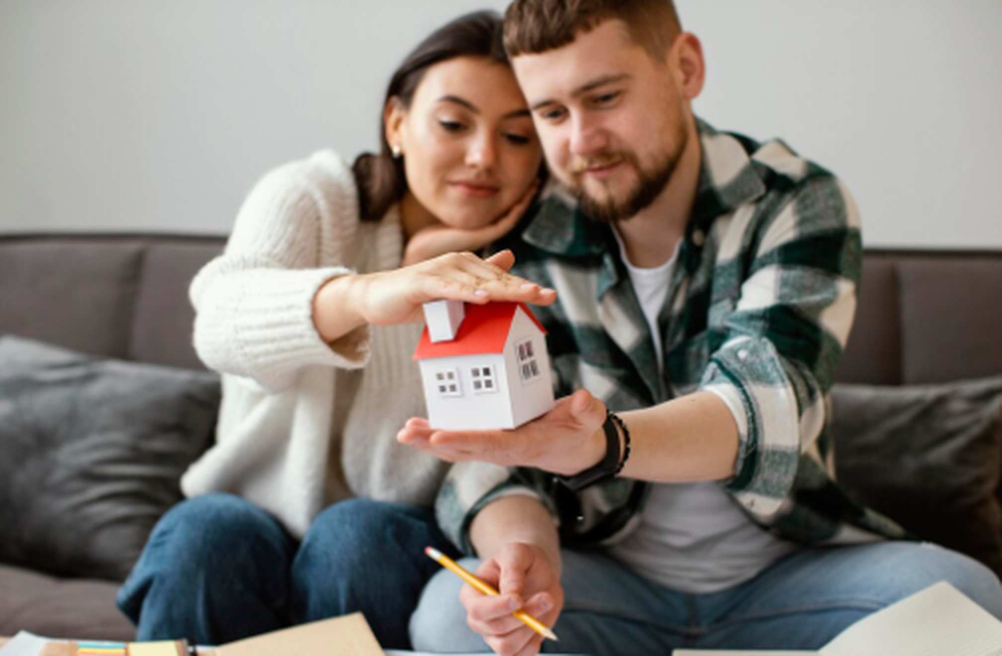 Casal jovem sentado no sofá segurando juntos uma pequena maquete de casa, representando o sonho de financiar um imóvel com planejamento e segurança.