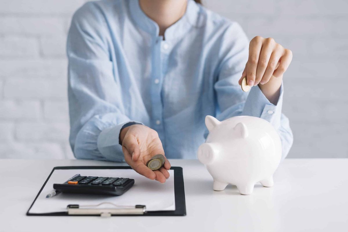 Pessoa colocando moedas em um cofrinho branco em formato de porco, ao lado de uma calculadora e documentos sobre a mesa, simbolizando economia, planejamento e cuidados com a saúde financeira.