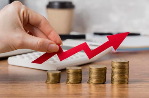 Mão posicionando uma seta vermelha em crescimento sobre pilhas de moedas, simbolizando o aumento dos custos financeiros e o impacto das taxas abusivas em empréstimo pessoal.