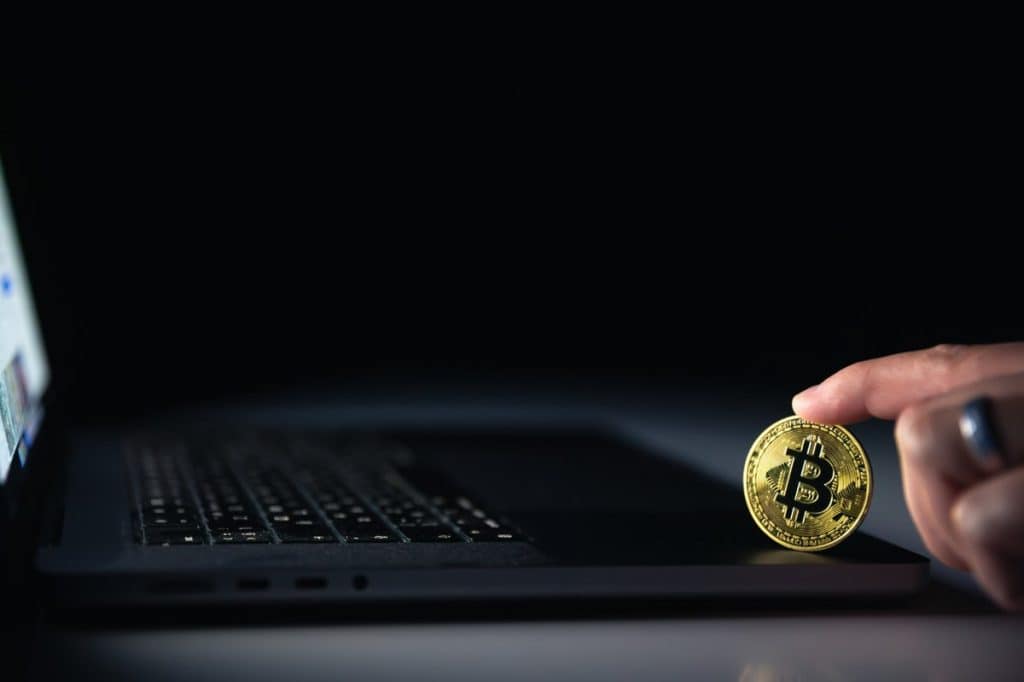 Mão segurando uma moeda física de Bitcoin dourada próxima a um laptop em ambiente escuro. A luz destaca o símbolo do Bitcoin, simbolizando empréstimo, investimento e transações digitais em criptomoedas.
