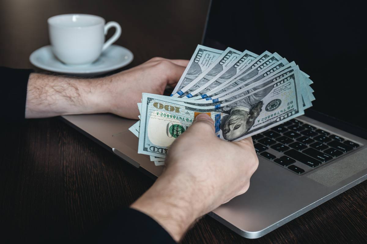 Mãos masculinas segurando notas de dólar americano sobre um laptop, com uma xícara de café ao fundo, representando novidades sobre ganhos financeiros, trabalho remoto e oportunidades de renda em dólares no cenário atual.